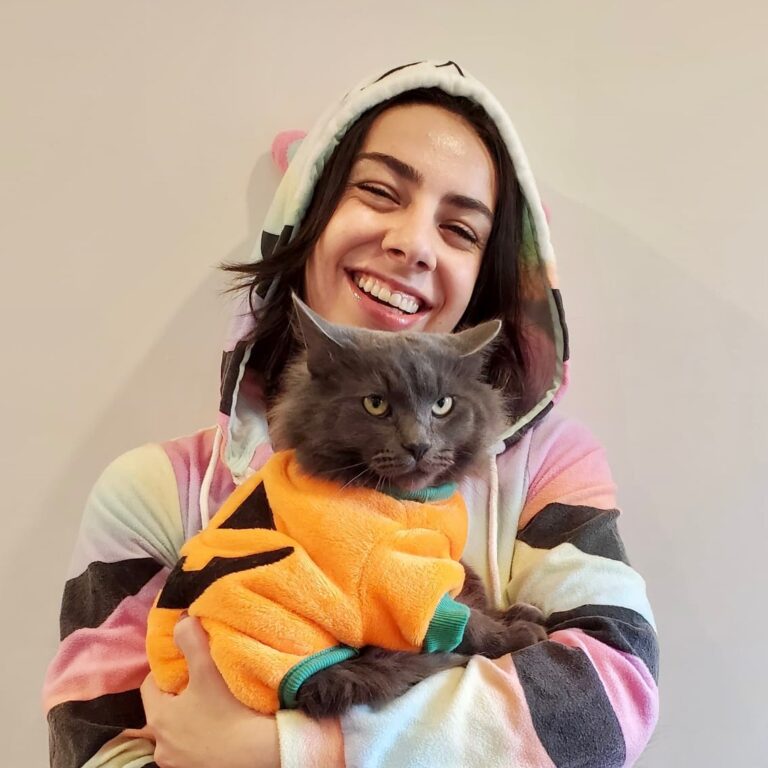 Woman wearing a Halloween cat costume while holding a cat dressed as a pumpkin